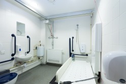 A larrger than average disabled toilet, with toilet, basin, ceiling hoist, shower ad adult sized changing table. White walls, dark blue grab rails.