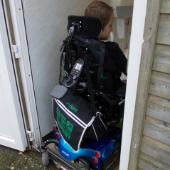 Boy using an adult sized powered wheelchair is unable to get in the disabled toilet with the whole of his chair. The rear castors are still outside the room. The chair is blue and the boy is visibly sad.