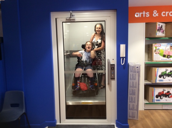 Wheelchair using child and his mum in a lift in a toy shop. The child is wearing blue shorts and a t-shirt shirt with Hogwarts Castle on it. Mum is wearing a black and white summer dress. The child is open mouthed and waving his arms in excitement. Mum is smiling widely.