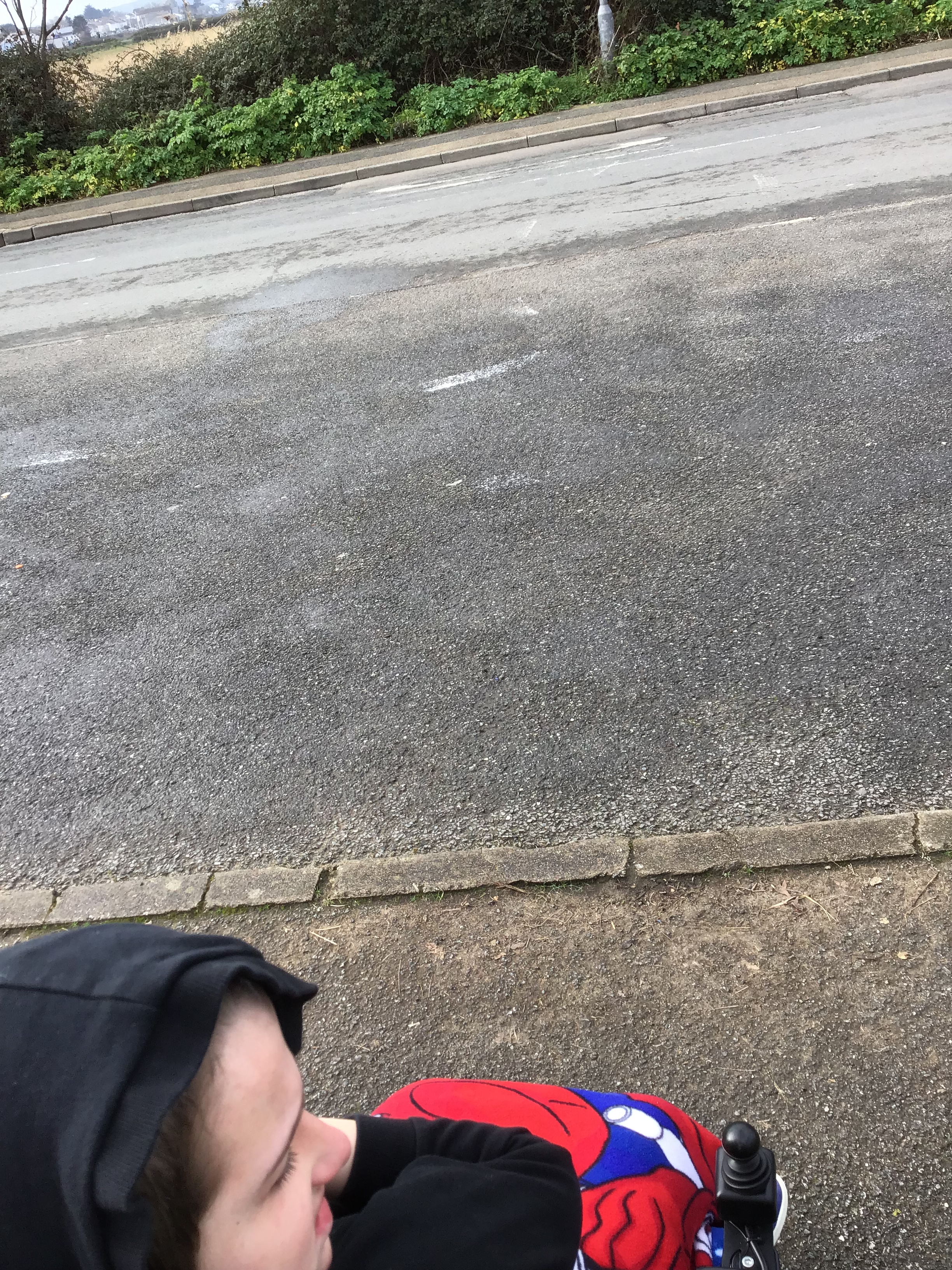 Photo of a road, with a lowered kerb on one side, but no lowered kerb on the other. A young male powered chair user is looking visibly sad.
