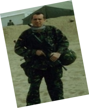 Man dressed in armed forces clothing in a sandy environment with an army tent in the background.