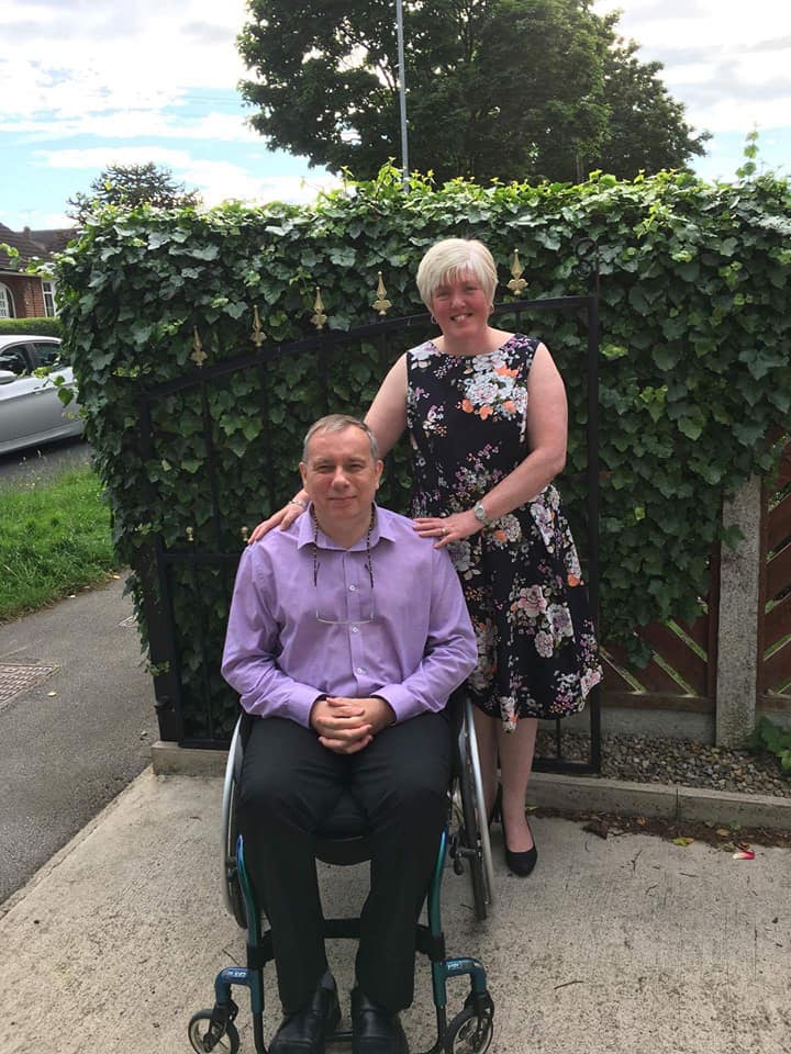 A smarltly dressed and smiling couple pose. The woman is stood behind the man, who is using a wheelchair. SHe is wearing a summery floral dressm the man is wearing black trousers and a lilac shirt.
