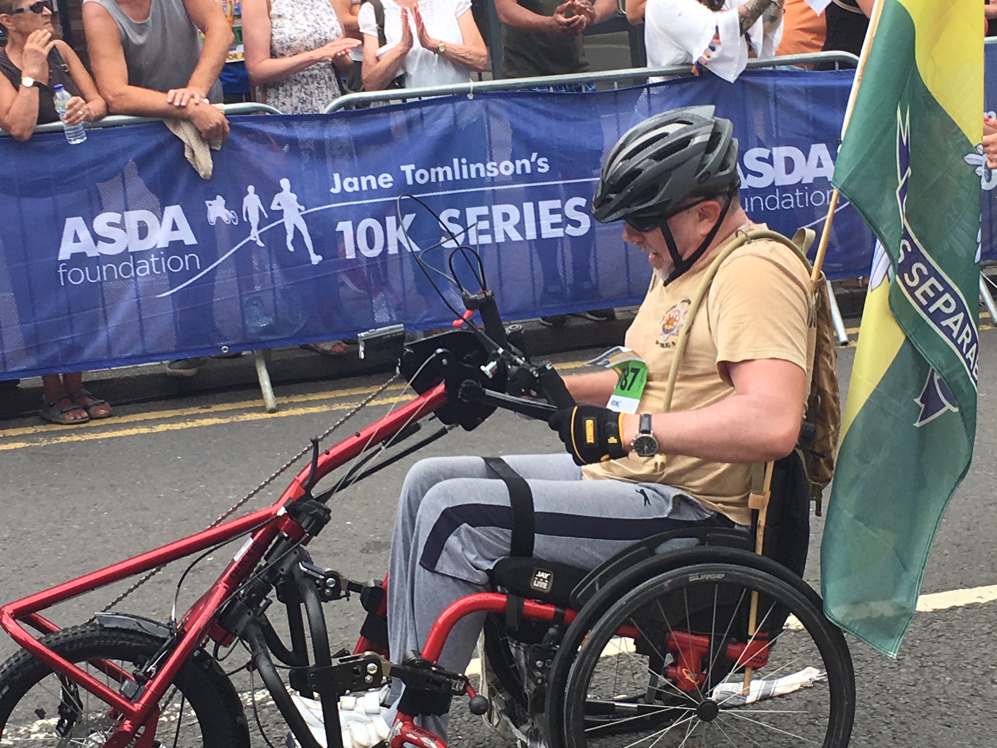 Man using a hand powered cycle. Man is wearing a grey cycling helmet, light coloured T-shirt and grea trousers. He looks determined. There is what appears to be a forces flag attached to the rear of his cycle. Signs behind him state "Asda Foundation Jane Tomlinson's 10K series.