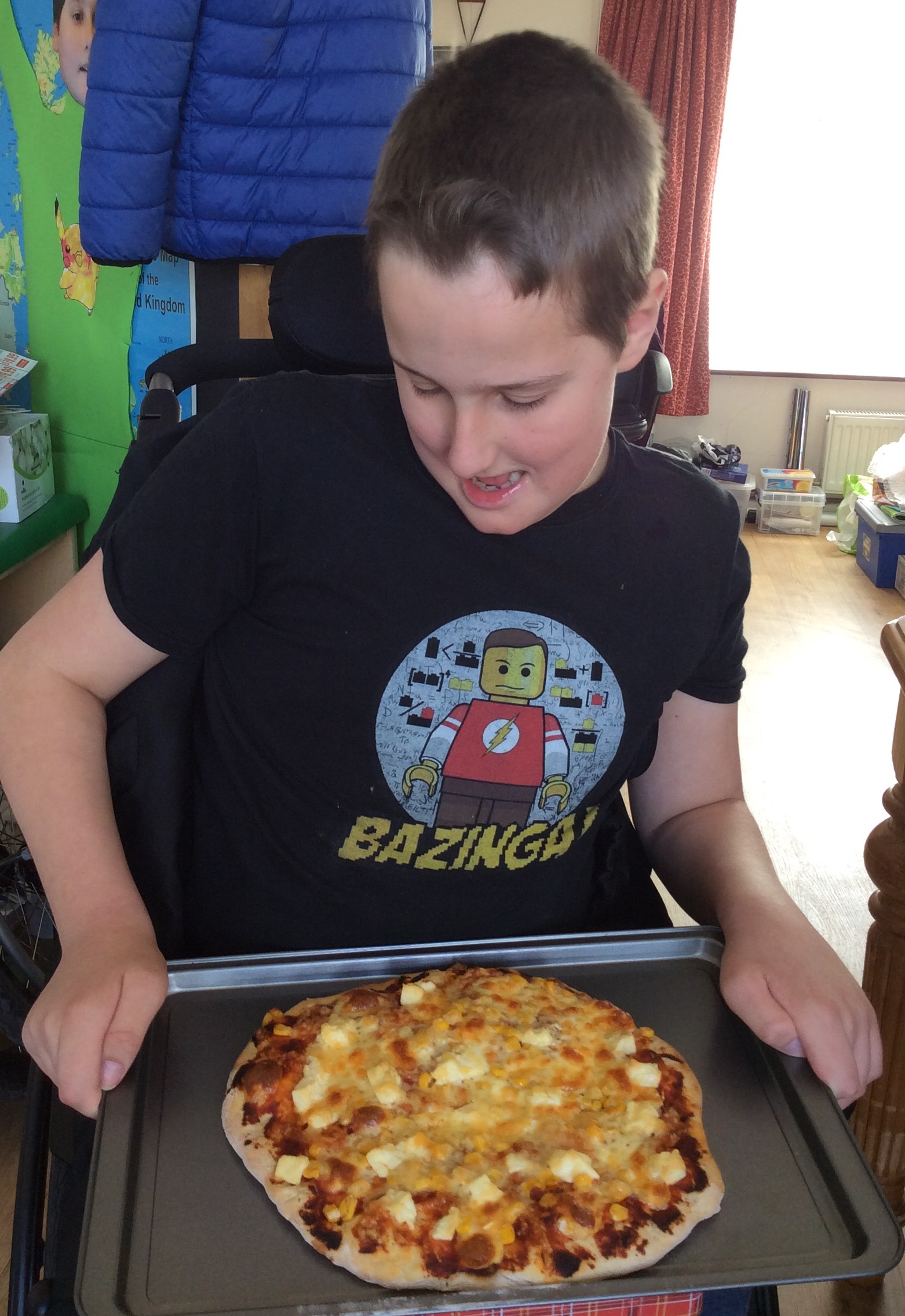 A boy using a wheelchair is smiling and looking down at his home made pizza. He is wearing a black Tshirt with a Lego Sheldon character on it and the word "Bazinga!"