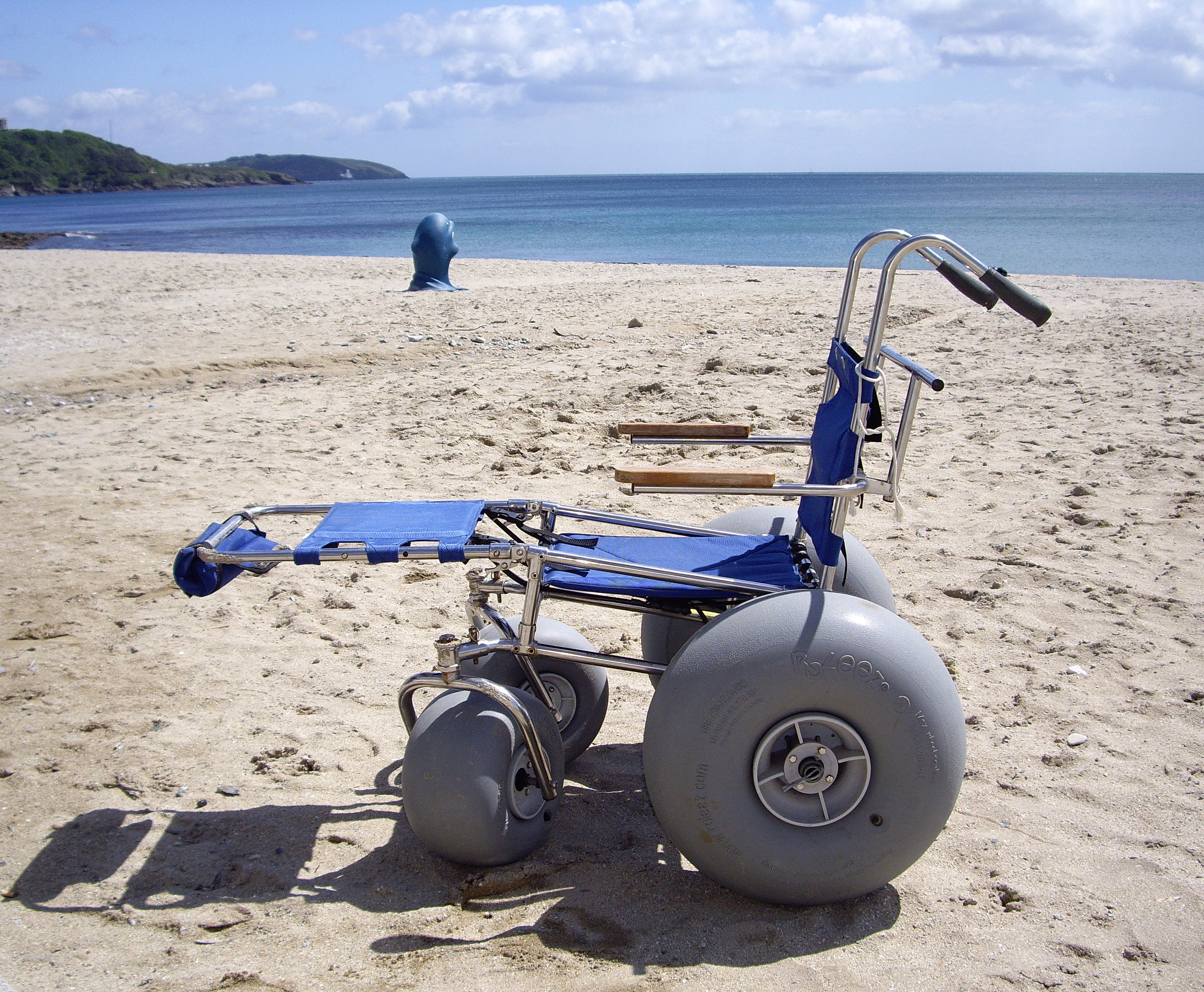 A sand chair on a beach, with the sea behind. It is a basic, upright mesh seatwith leg support extended. Woor armrests on metal poles, no head support, no body support and no lap belt or supportive harness. Large air wheels at the front and huge air wheels at the rear.